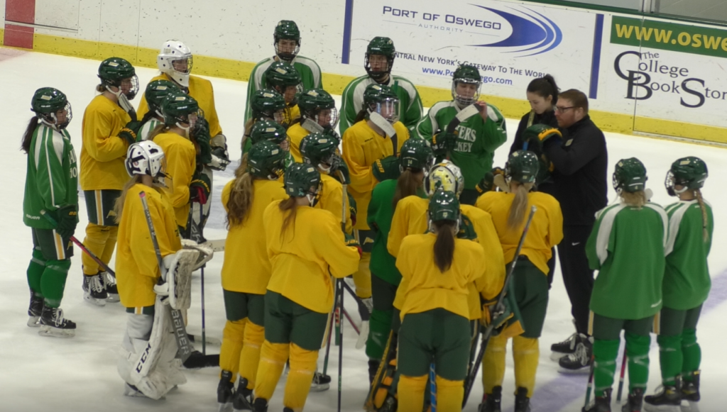The Oswego State women's hockey roster forming around their coach on the rink in Marano Campus Center