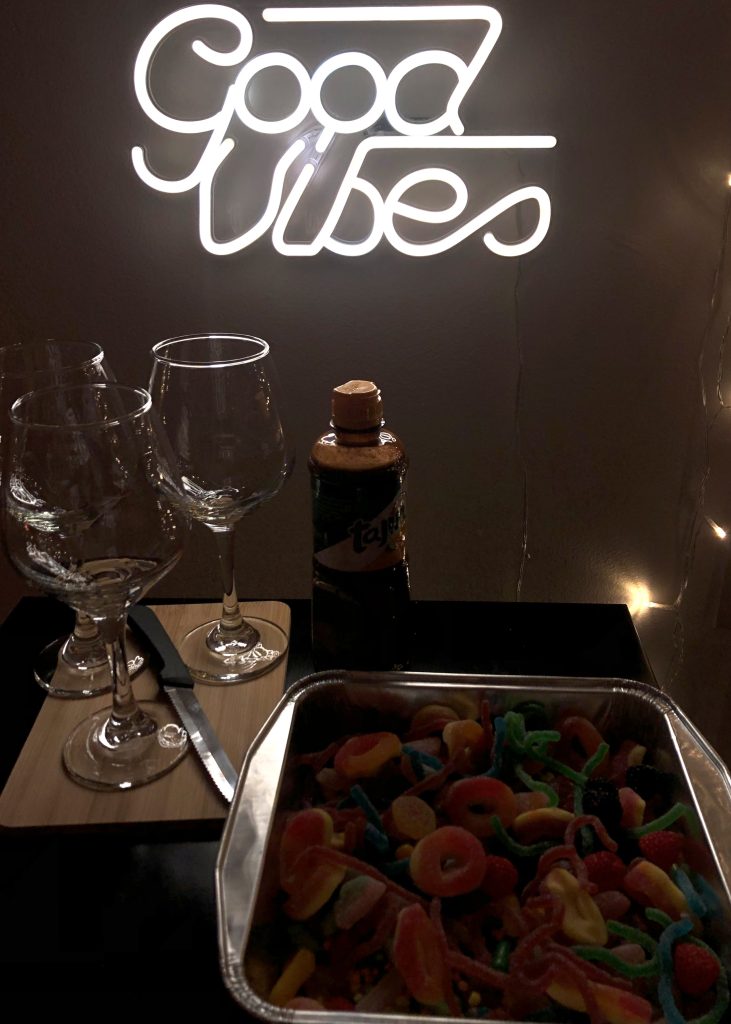 A bowl of mixed candy, a bottle of Chamoy, and glass cups sit on a counter with a glowing sign behind them saying "good vibes."