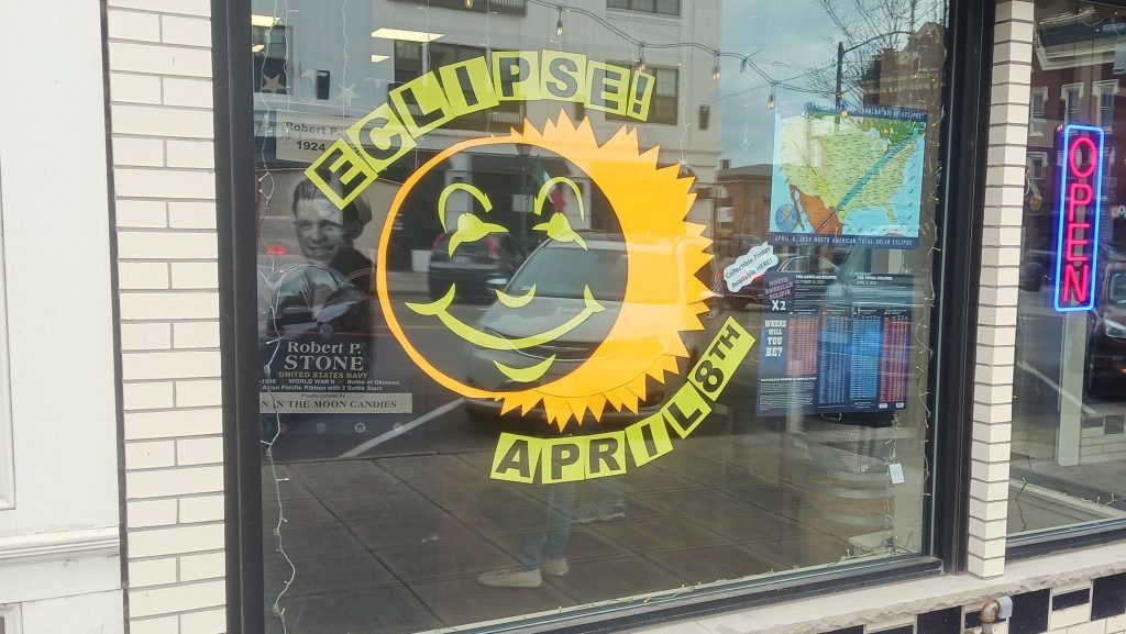 A window display shows a smiling moon on the sun, emulating a solar eclipse. Text around decal reads, "Eclipse! April 8th."